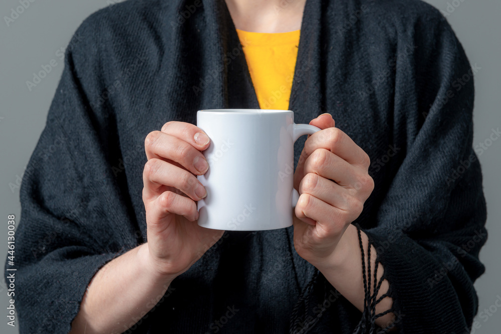 woman in a black cloak is holding white mug with hot drink in both hands woman in a black cloak is holding white mug with hot drink in both hands