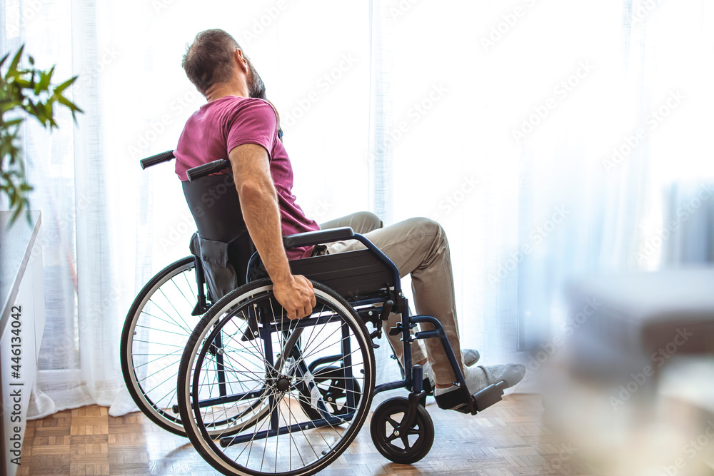 Obraz premium Side view of mature man sitting in wheelchair and looking away. Side view of thoughtful disabled active Caucasian man looking through window on wheelchair in a comfortable home