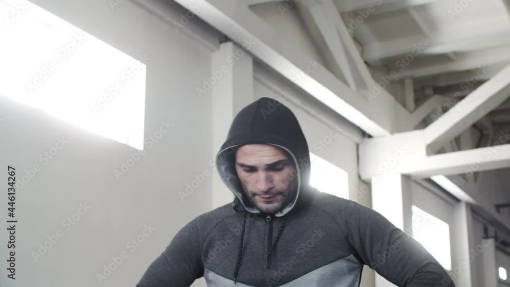 Pensive young sportsman resting and thinking in gym. Low angle view of thoughtful athletic man in sportswear and hood walking and looking down in gym Pensive young sportsman resting and thinking in gym. Low angle view of thoughtful athletic man in sportswear and hood walking and looking down in gym