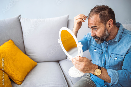 Middle-aged man concerned by hair loss. Senior man and hair loss issue. Middle aged man with alopecia looking at mirror, hair loss concept. Bearded mid adult man with alopecia looking at mirror photo