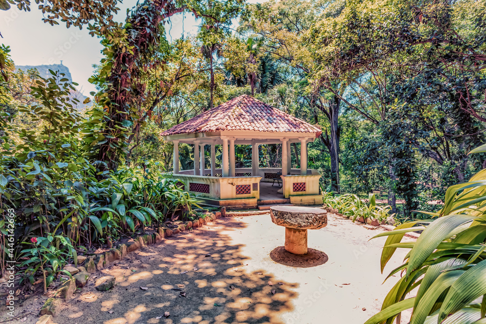 The exterior of a gazebo structure on a paved path in a tropical garden on a sunny day.
