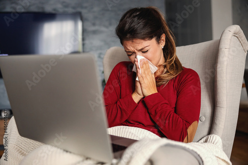 Sick woman sitting in chair at home during lockdown with laptop in her lap and blowing nose.