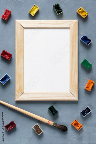 Stationery mock-up scene. Blank white greeting card in wooden frame surrounded bywatercolor paints in ditches and a round squirrel brush on textured gray background.