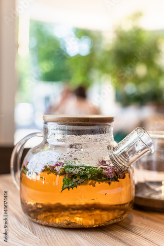 Green or herbal tea in glass teapot with mint and roses petals on wooden table in cafe. Blurred background.