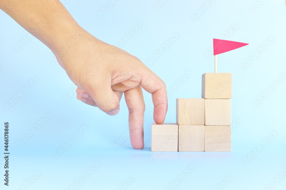 Human hand fingers climbing stairs of wooden blocks with red flag. Take first step on ladder to success and goal. Human hand fingers climbing stairs of wooden blocks with red flag. Take first step on ladder to success and goal.