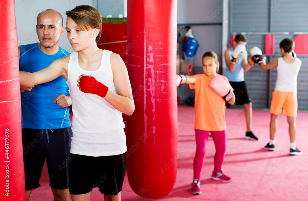 Adult boxing instructor and glad young children practicing blows on boxing bag