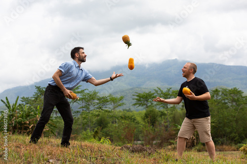 Two caucasian hipsters play with papaya and pineapple outdoors, friend, joke, vegetarian