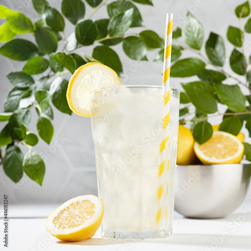 Fresh natural healthy vitamin citrus drink in glass with ice, straw, yellow lemons in metallic bowl, green foliage in soft light white kitchen with marble tile on white wood table, square, closeup.