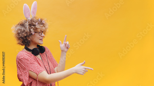 Attractive young girl wearing cute bunny ears and spectacles in a joyful mood. Headphones around her neck. Girl smiling and pointing at something. Bright yellow background. Studio shot. photo