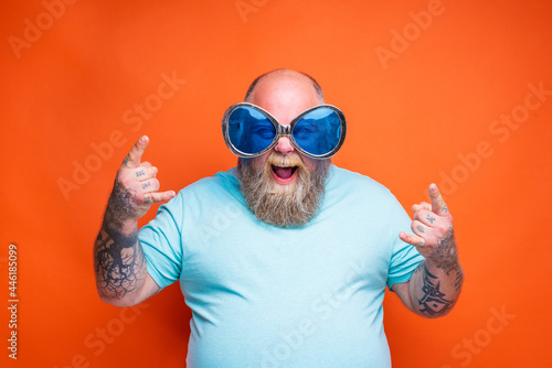 Fat happy man with beard, tattoos and sunglasses makes the gesture of the horns photo