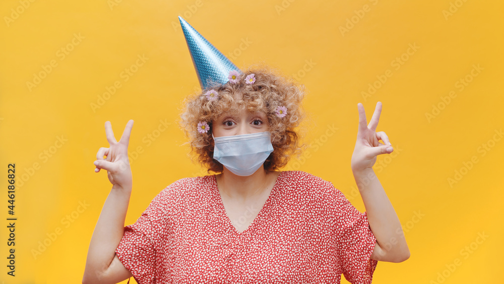 Girl wearing a birthday cap and a disposable face mask. Making victory signs. Dressed in a pink shirt, wearing flowers in her hair. Girl in a festive mood. Concept of Covid19 and social distancing. Girl wearing a birthday cap and a disposable face mask. Making victory signs. Dressed in a pink shirt, wearing flowers in her hair. Girl in a festive mood. Concept of Covid19 and social distancing.