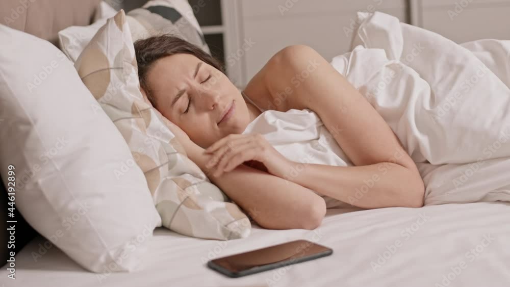 Medium close-up of young Caucasian woman lying on her side in bed, waking up in morning, looking at smartphone, frowning and getting back asleep Medium close-up of young Caucasian woman lying on her side in bed, waking up in morning, looking at smartphone, frowning and getting back asleep