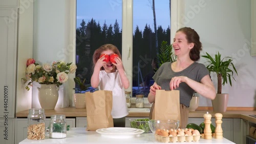 Disassemble food in the kitchen from paper bags. Mom and daughter play and laugh together at home. Have fun with home delivery. Dance and play with vegetables and fruits for breakfast or dinner.