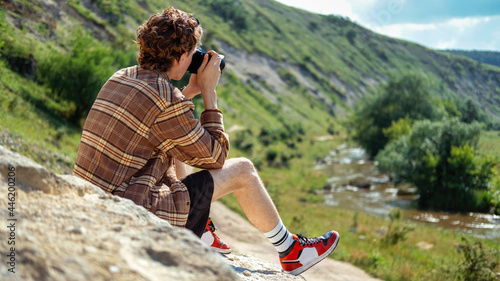 Obraz na płótnie A young man with curly hair taking shots using camera in the nature, sitting on