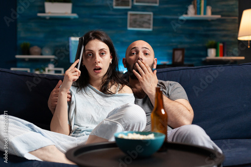 Astonished couple watching movie at night and eating popcorn, drinking beer reacting at film emotion. Focused concentrated confused wife chilling home on confortable couch having facial expression