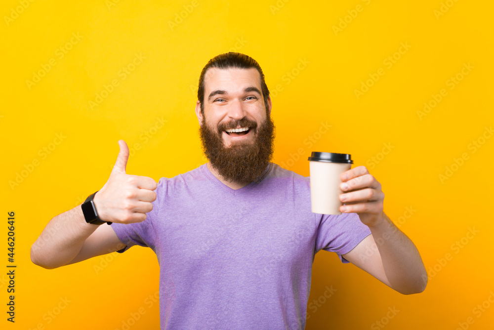 Young smiling bearded drinking hold paper cup of coffee show thumb up gesture isolated on yellow background Young smiling bearded drinking hold paper cup of coffee show thumb up gesture isolated on yellow background
