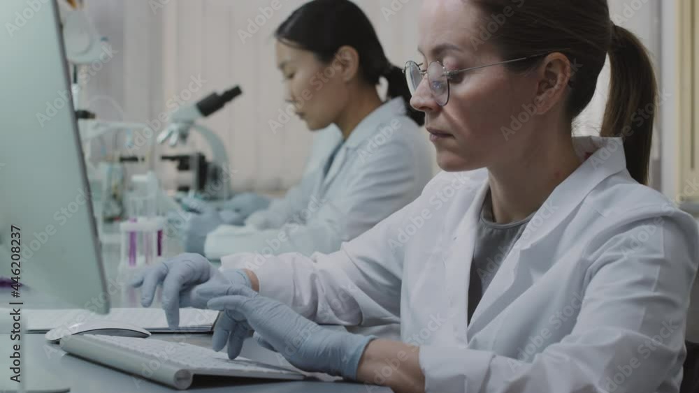 Tilt up slowmo shot of young female scientist in white coat, gloves and glasses typing on computer in laboratory while her male and female colleagues working in background Tilt up slowmo shot of young female scientist in white coat, gloves and glasses typing on computer in laboratory while her male and female colleagues working in background