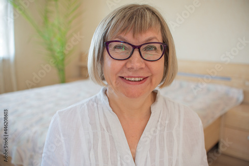 Happy senior woman with gray hair with glasses relaxing smiling is looking at the camera in her home.
