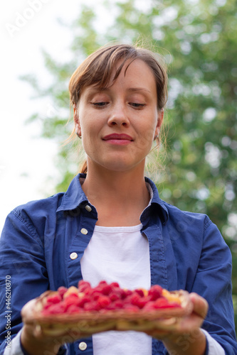 A beautiful young woman in a blue shirt holds a basket of fresh fragrant raspberries in her hands against a background of green foliage. Selective focus. Portrait
