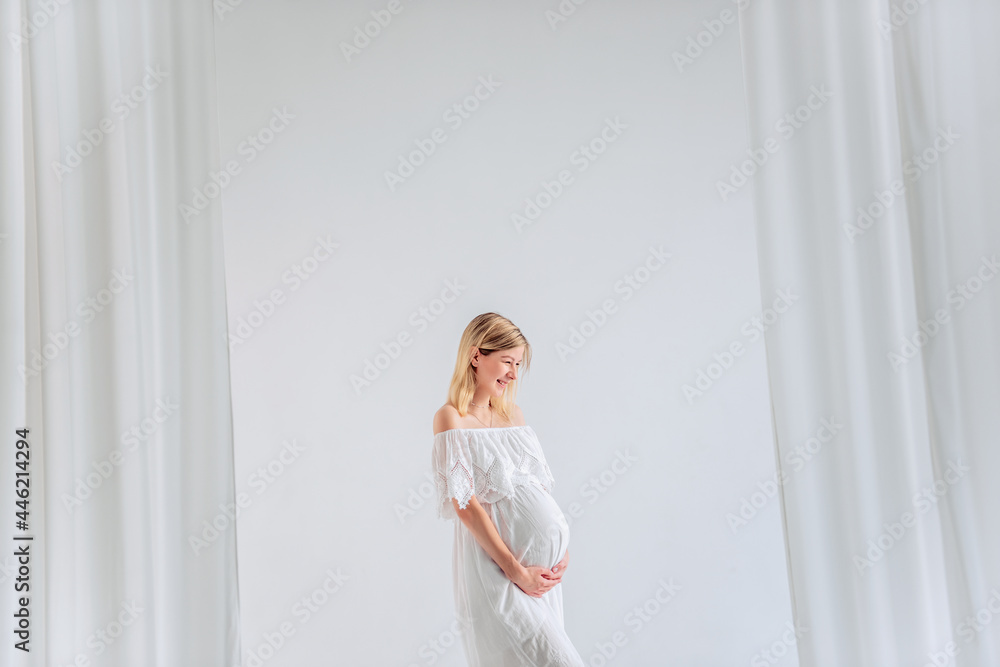 A young pregnant woman in anticipation of a baby in a white dress, hugs belly. Fashion girl in a minimalistic, isolated interior of a cut of airy fabrics. Easy, happy maternity pregnancy, surrogacy. A young pregnant woman in anticipation of a baby in a white dress, hugs belly. Fashion girl in a minimalistic, isolated interior of a cut of airy fabrics. Easy, happy maternity pregnancy, surrogacy.