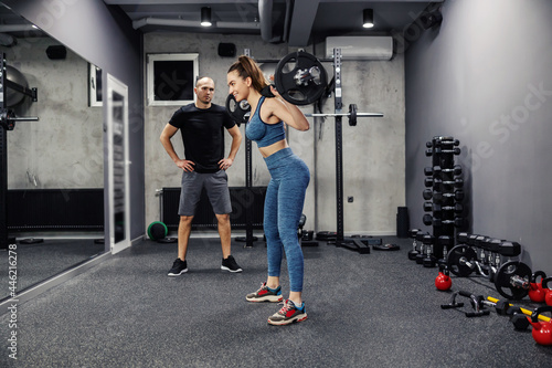 Barbell training with weights. A young woman, in sportswear and in good shape, does squats and holds a barbell on her back. In the background is an individual fitness trainer who is support her © dusanpetkovic1