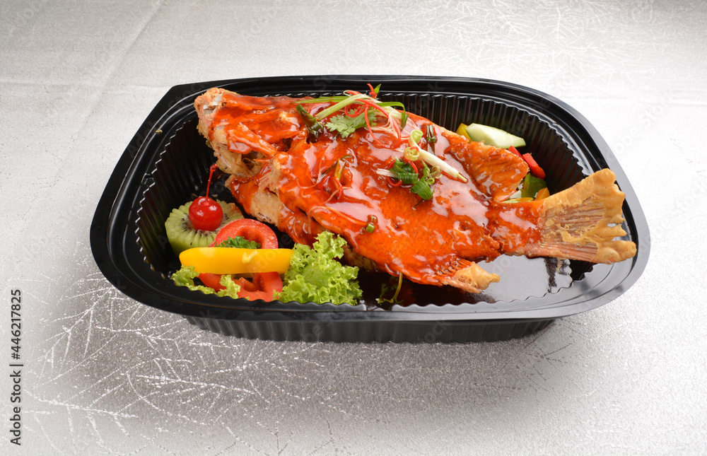 deep fried crispy whole grouper fish seafood with sweet and sour chilli sauce and lychee fruits on top in black bento tray box for asian take away halal menu