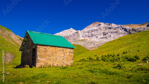 Paisaje de montaña en el que se aprecia un refugio en primer plano rodeado de pastos y montañas de fondo con un cielo azul