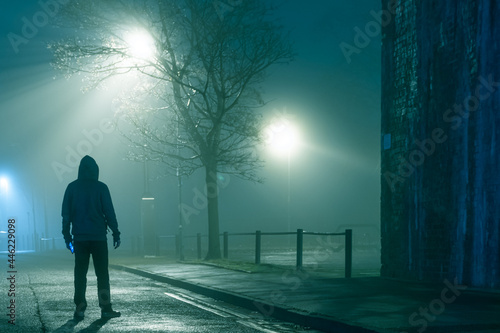 A mysterious hooded figure holding a phone  standing in a city street. On a moody foggy winters night