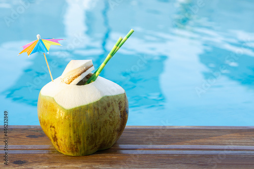 Fresh  green coconut drink with paper straw  and rainbow umbrella  standing on wooden board  near pool  water  tropical beach resort background  with copy space Vacation  in exotic  resort  concept .