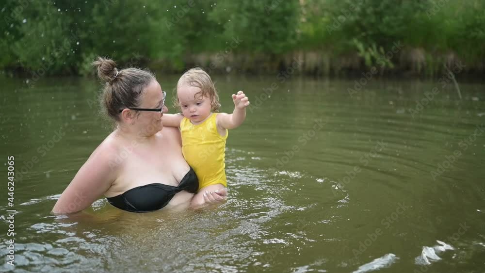 Little funny cute blonde girl child toddler in yellow bodysuit laughing learns swim outside at summer lake. Plus size body positive smiling woman mother baby swimming in natural pool. Water splashes Little funny cute blonde girl child toddler in yellow bodysuit laughing learns swim outside at summer lake. Plus size body positive smiling woman mother baby swimming in natural pool. Water splashes