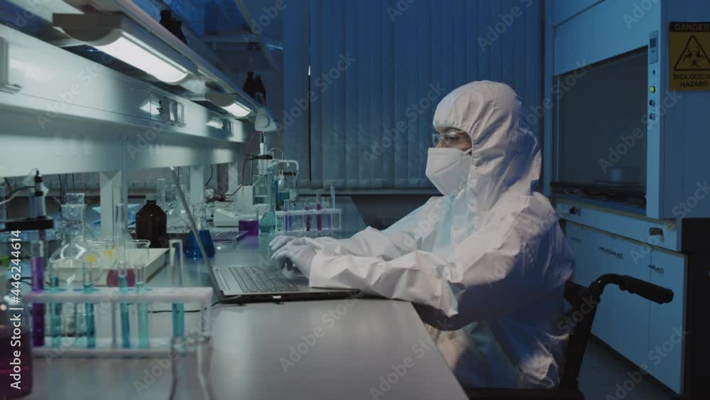 PAN slowmo shot of unrecognizable female epidemiologist in disposable coveralls, googles, face mask and gloves sitting in wheelchair and working on laptop in laboratory, then looking at camera PAN slowmo shot of unrecognizable female epidemiologist in disposable coveralls, googles, face mask and gloves sitting in wheelchair and working on laptop in laboratory, then looking at camera