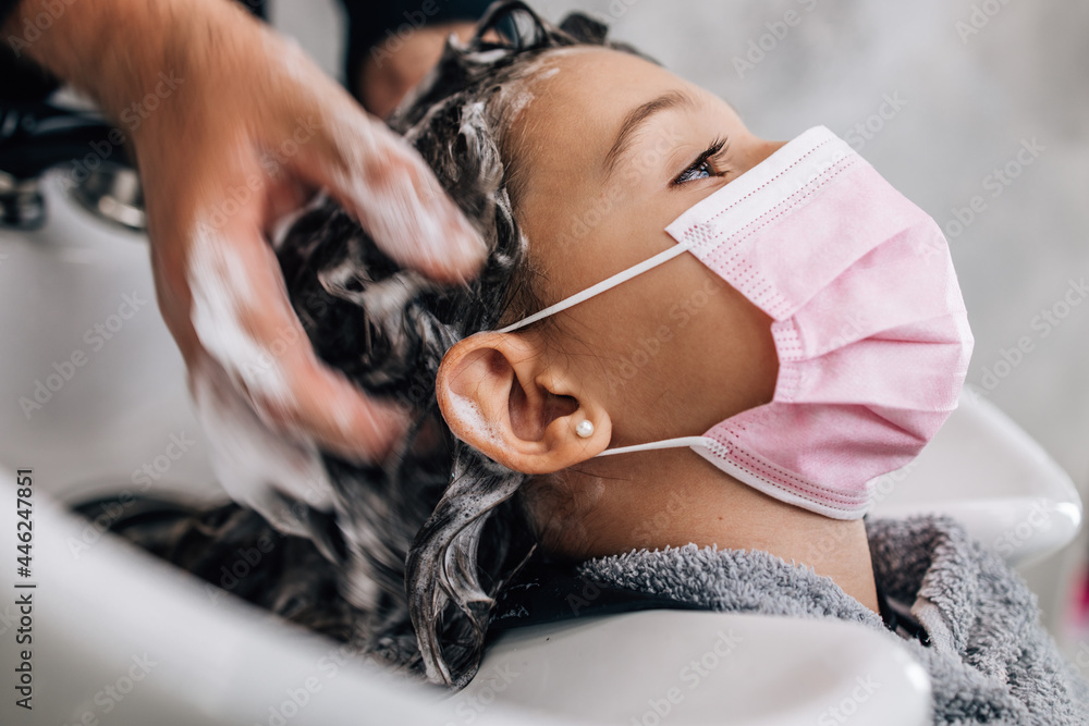 Young girl enjoying at hairstyle treatment while professional hairdresser gently working and cutting her hair. They are wearing protective face mask against virus pandemic