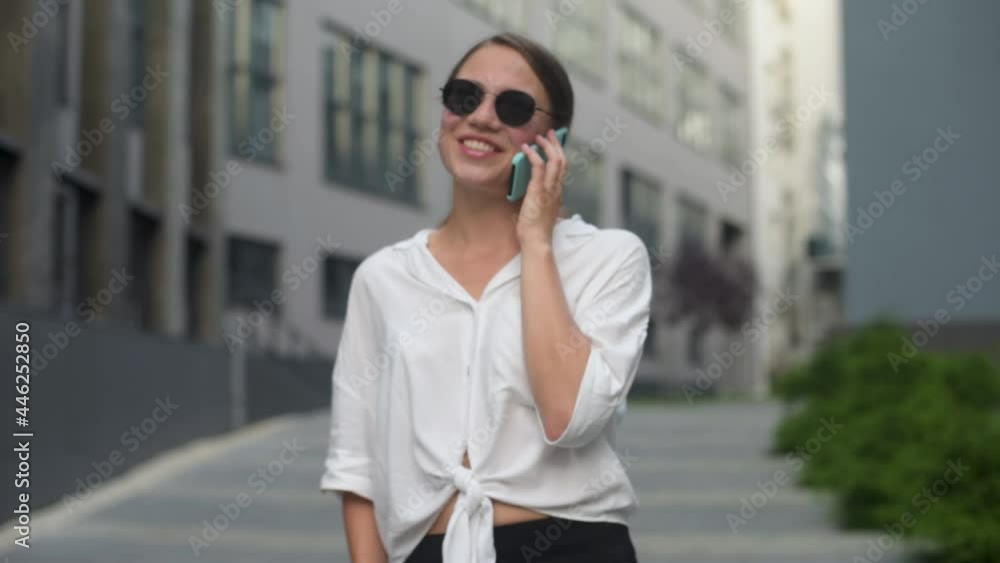 Smiling lady holding cellphone in hands outside. Business woman wearing trendy sunglasses walks down the central city street and uses her phone. Smiling lady holding cellphone in hands outside. Business woman wearing trendy sunglasses walks down the central city street and uses her phone.