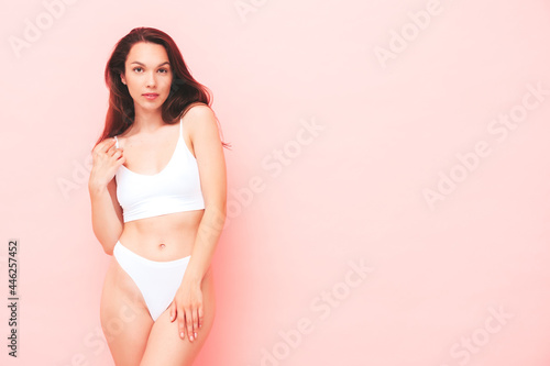 Portrait of young beautiful smiling woman in white lingerie. Sexy carefree cheerful model in underwear posing near pink wall in studio. Positive and happy female enjoying morning