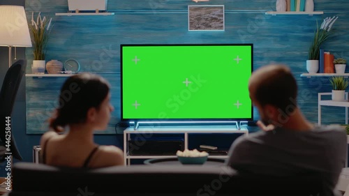 Caucasian man and woman looking at green screen on television display at home sitting in living room. Young couple with copy space on modern chroma key for isolated mockup template photo