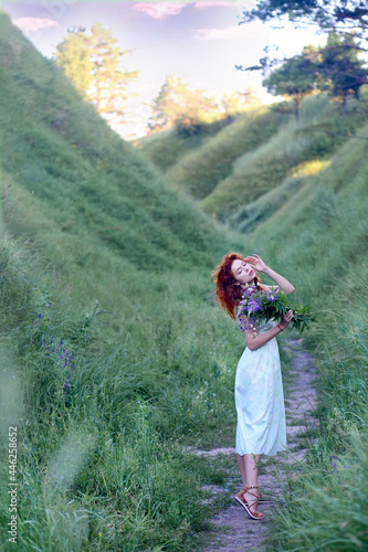 A young beautiful red-haired girl in a green sundress in the summer walks where the hills and mountains  green grass and wildflowers.