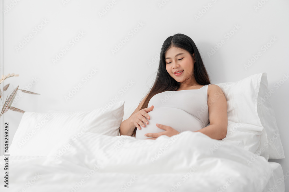 Asian happy pregnant woman is sitting on bed and and touching her belly. pregnancy, motherhood, people and expectation concept Asian happy pregnant woman is sitting on bed and and touching her belly. pregnancy, motherhood, people and expectation concept