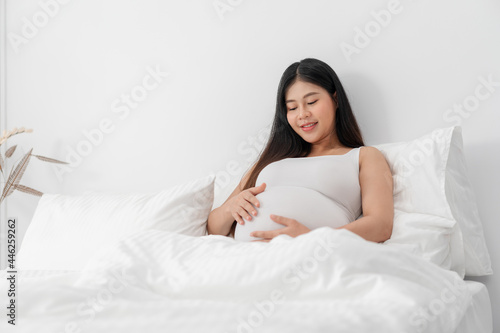 Asian happy pregnant woman is sitting on bed and and touching her belly. pregnancy, motherhood, people and expectation concept