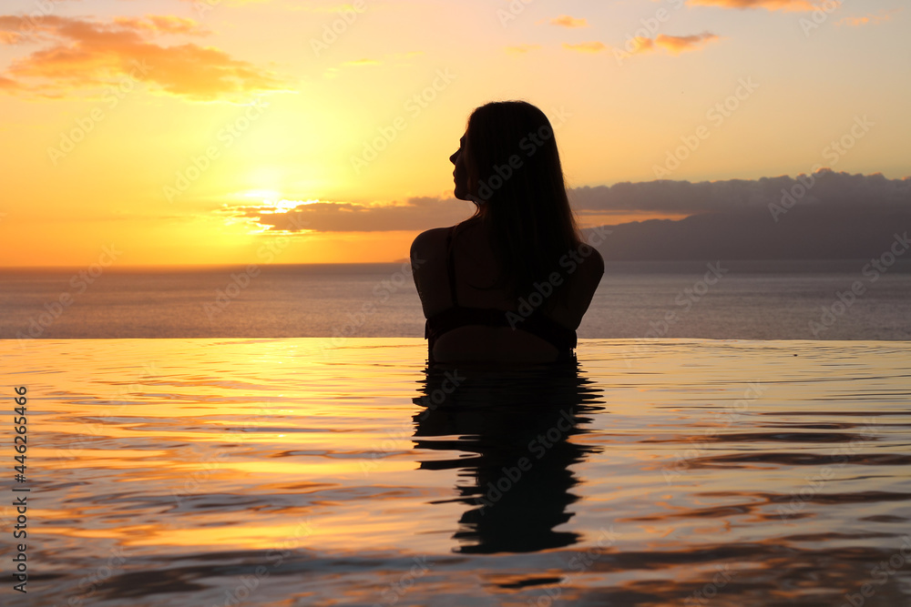  A girl in a swimsuit with her hair down stands back in the pool and looks at the sunset and the ocean.