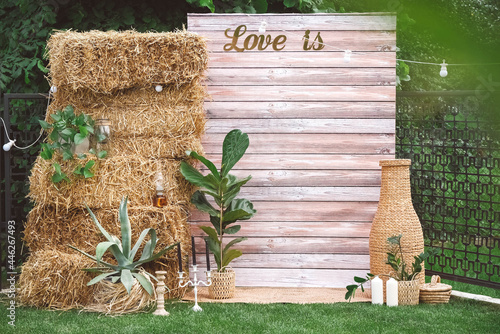 Rustic wedding photo zone decorated with wooden background, hay, wicker pots with plants, lamps and candles. Copy, empty space for text
