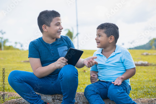 Latino brothers talking while looking at the tablet sitting on the tree trunk in a field photo