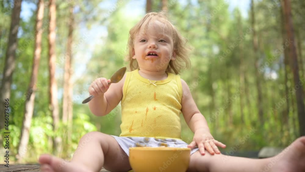 Little funny cute blonde girl child toddler with dirty clothes and face eating baby food fruit or vegetable puree with spoon from yellow plate outside at summer. Healthy happy childhood concept Little funny cute blonde girl child toddler with dirty clothes and face eating baby food fruit or vegetable puree with spoon from yellow plate outside at summer. Healthy happy childhood concept