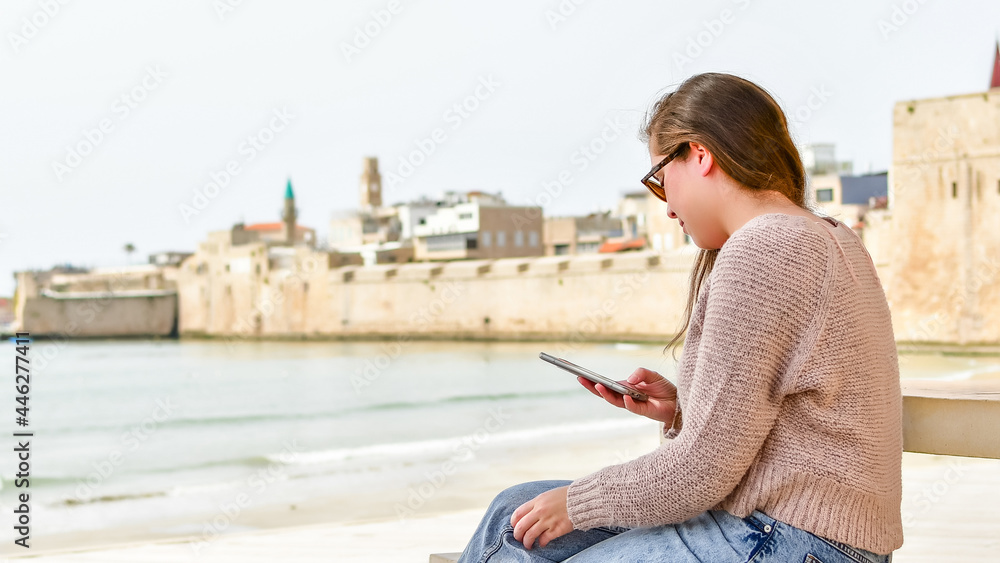 Side view of young white woman, using her mobile phone, sitting on a mediterranean beach in the Middle East.