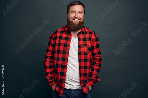 Good-looking stylish modern hipster guy with stylish haircut, beard, wear red checkered shirt, hold hands in pockets and smiling joyfully, laughing chatting with friends, grey background
