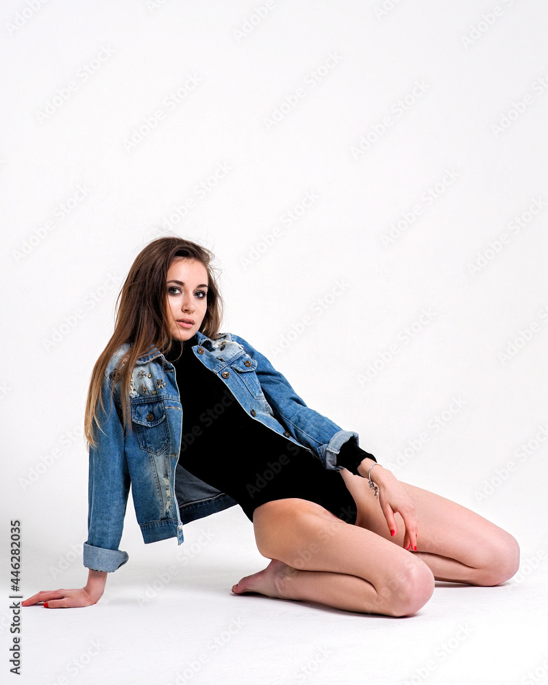 Fototapeta premium The brunette is on her knees on a white background in a bodysuit and a denim jacket