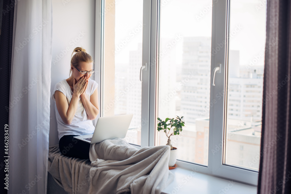 Happy young adult woman freelancer wearing glasses sitting on window background. Laughing blonde working on laptop. Workplace on the windowsill. Home office concept. Happy young adult woman freelancer wearing glasses sitting on window background. Laughing blonde working on laptop. Workplace on the windowsill. Home office concept.