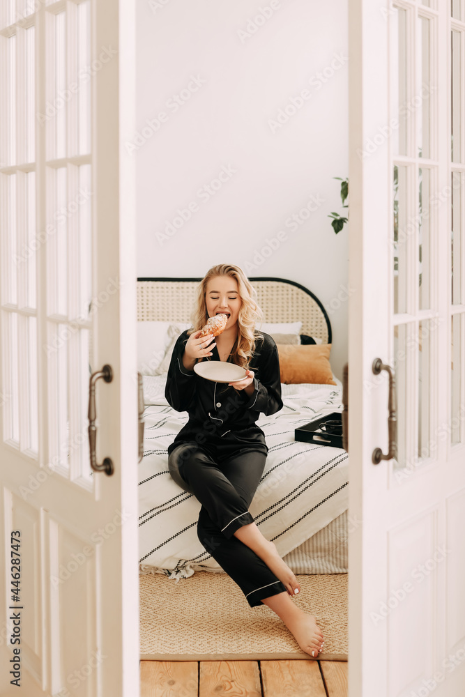 A cheerful happy sleepy young woman in black comfortable pajamas alone enjoys relaxing having breakfast drinking coffee in bed in a bright bedroom in a cozy house on a weekend morning A cheerful happy sleepy young woman in black comfortable pajamas alone enjoys relaxing having breakfast drinking coffee in bed in a bright bedroom in a cozy house on a weekend morning