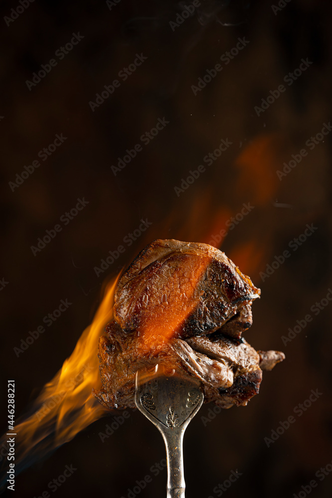 Tasty beef steak on a fork with fire flames, smoke and salt on dark background. Freeze motion barbecue concept. Close up macro view on meat, copy space. Juicy grilled piece of meat. Tasty beef steak on a fork with fire flames, smoke and salt on dark background. Freeze motion barbecue concept. Close up macro view on meat, copy space. Juicy grilled piece of meat.