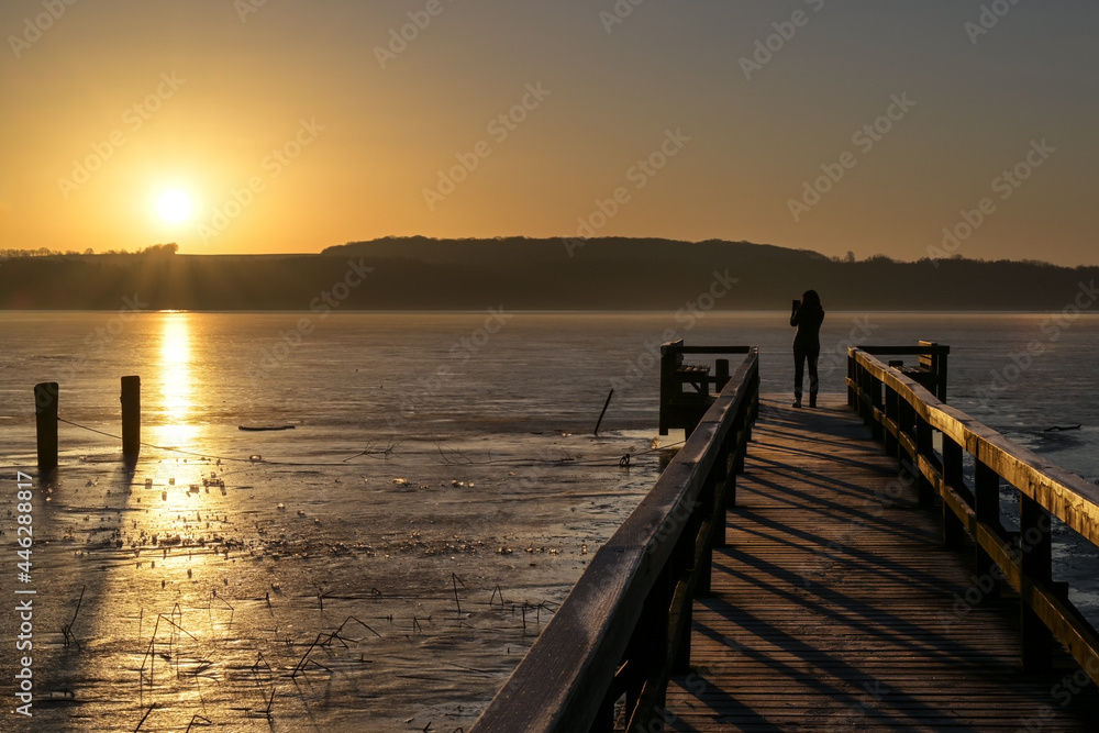 Young woman from behind as silhouette on a wooden jetty taking a photo of the rising sun over the frozen lake, copy space