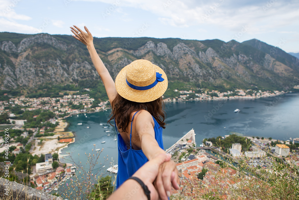 A girl in a blue dress and a straw hat held by a musky hand stands against the background of Boka Kotorska Bay on a beautiful summer day, Montenegro.